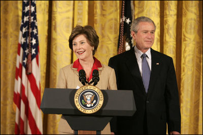 President George W. Bush listens on as Mrs. Laura Bush welcomes women leaders to the East Room for a celebration Tuesday, March 7, 2006, of International Women's Day. Mrs. Bush said, "I've been privileged to meet thousands of women from many nations, and I believe that women everywhere share the same dreams -- to be educated, to live in peace, to enjoy good health, to be prosperous, and to be heard."