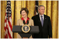 President George W. Bush listens on as Mrs. Laura Bush welcomes women leaders to the East Room for a celebration Tuesday, March 7, 2006, of International Women's Day. Mrs. Bush said, "I've been privileged to meet thousands of women from many nations, and I believe that women everywhere share the same dreams -- to be educated, to live in peace, to enjoy good health, to be prosperous, and to be heard."