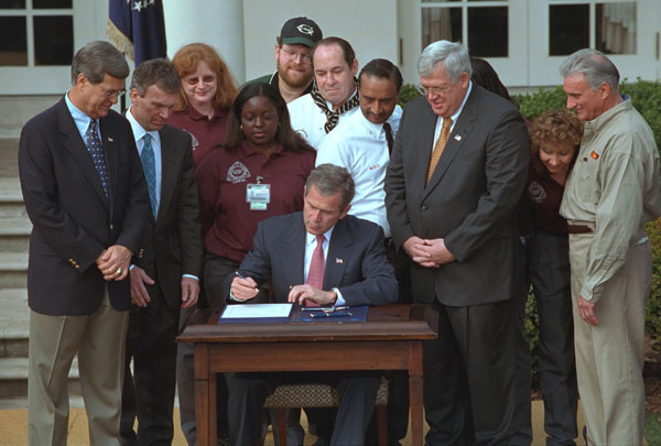 President George W. Bush signs the Job Creation and Worker Assistance Act of 2002 during a live radio address in the Rose Garden March 9, 2002. On hand for the signing were Senators Trent Lott (far left) and Tom Daschle (second from left) and Speaker of the House Dennis Hastert (standing to right of the President). "We're seeing some encouraging signs in the economy, but we can't stand by and simply hope for continued recovery," said President Bush. "We must work for it." White House photo by Eric Draper.