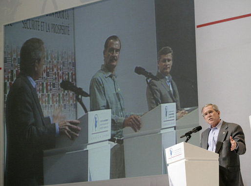 President George W. Bush addresses the media as Mexico's President Vicente Fox and Canadian Prime Minister Stephen Harper are seen on a large video screen listening during their joint news conference, Friday, March 31, 2006 at the Fiesta Americana Condesa Cancun Hotel in Cancun, Mexico. White House photo by Eric Draper